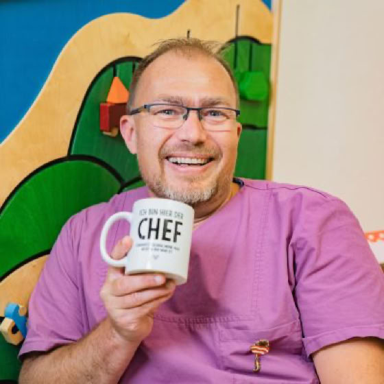 Ein lächelnder Mann mit Brille hält eine Tasse mit der Aufschrift "CHEF" vor einem bunten Hintergrund.