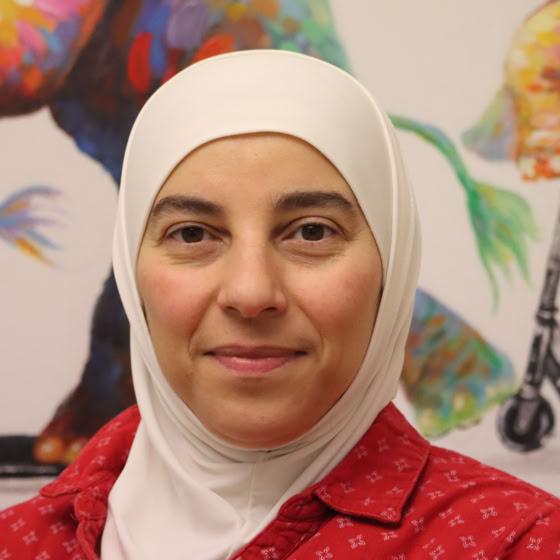 Eine Person mit einem weißen Kopftuch und einem roten Hemd steht vor einem bunten Hintergrund.